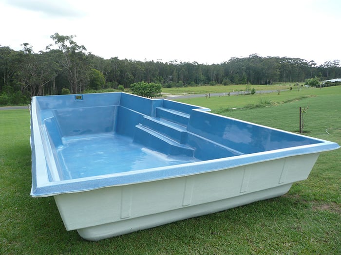 A large light blue fibreglass pool shell with built-in steps and bench seat sitting on green grass in a semi-rural Australian setting, showing the complete factory-moulded product